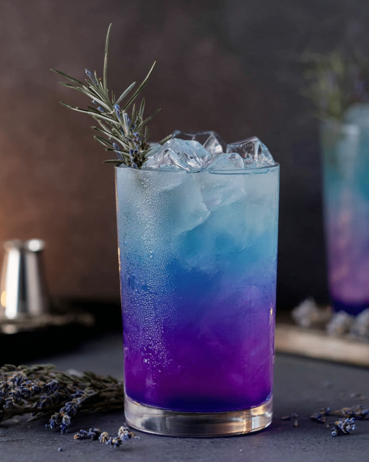 Chilled glass of Witches Brew Lemonade with layered purple and blue hues, garnished with a sprig of rosemary and served over ice.