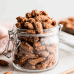 Glass jar filled with crunchy candied almonds coated in a cinnamon-sugar mixture, with a few almonds scattered around.