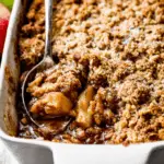 Homemade apple crisp with a golden oat topping, served warm in a white baking dish with a spoon.