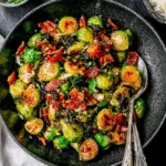 A bowl of roasted Brussels sprouts with crispy bacon pieces, garnished with herbs and served with a spoon.