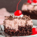 Slice of Cherry Dr Pepper Cake topped with cherry whipped frosting, chocolate shavings, and a maraschino cherry.