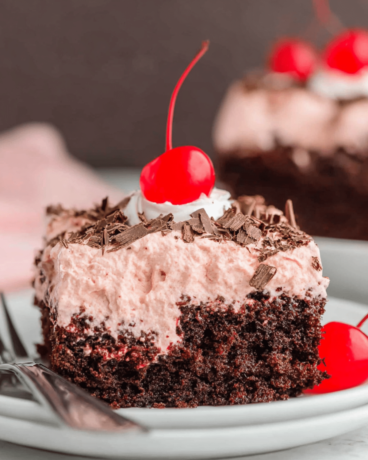 Slice of Cherry Dr Pepper Cake topped with cherry whipped frosting, chocolate shavings, and a maraschino cherry.