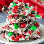 Stacked pieces of Christmas candy bark made with white chocolate, red and green candies, chocolate chips, and crushed peppermint on a white plate.