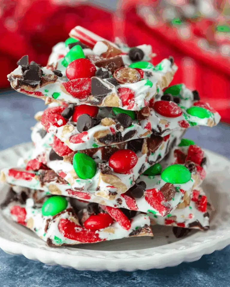 Stacked pieces of Christmas candy bark made with white chocolate, red and green candies, chocolate chips, and crushed peppermint on a white plate.