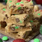 Stacked Christmas cookie bars filled with chocolate chips and red and green M&M candies, surrounded by festive holiday ornaments.