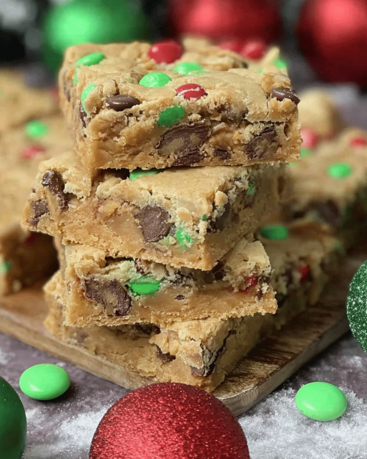 Stacked Christmas cookie bars filled with chocolate chips and red and green M&M candies, surrounded by festive holiday ornaments.