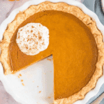 Classic pumpkin pie in a white pie dish with a slice missing, topped with whipped cream and cinnamon.