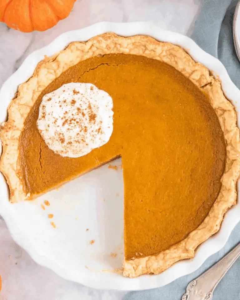 Classic pumpkin pie in a white pie dish with a slice missing, topped with whipped cream and cinnamon.