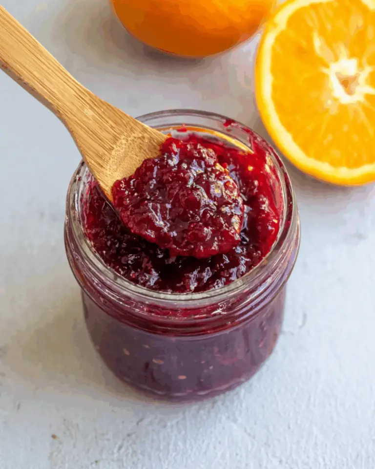 Jar of cranberry-orange preserves with a wooden spoon, surrounded by fresh orange halves.