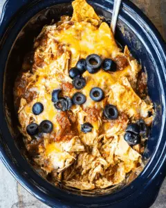 Crockpot chicken enchilada casserole with shredded chicken, melted cheese, salsa, tortilla chips, and sliced black olives.