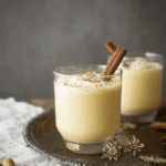 A glass of creamy eggnog garnished with ground nutmeg and a cinnamon stick, placed on a decorative tray with another glass of eggnog in the background.