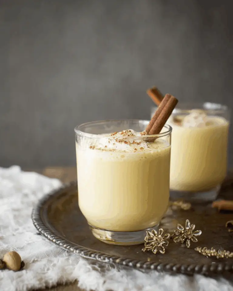 A glass of creamy eggnog garnished with ground nutmeg and a cinnamon stick, placed on a decorative tray with another glass of eggnog in the background.