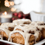 A plate of festive cinnamon rolls topped with rich vanilla icing, set against a cozy holiday background with lights and candles.