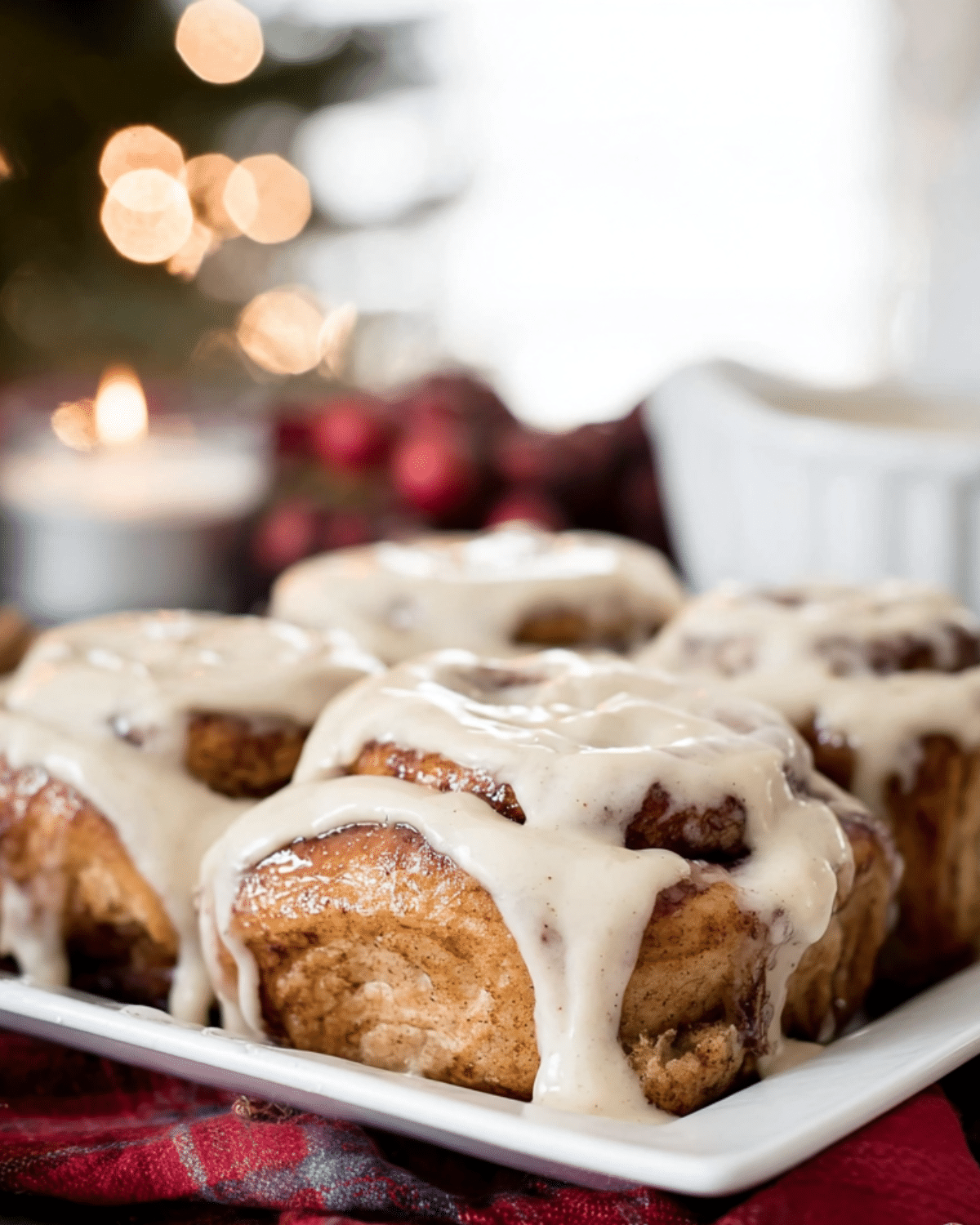 A plate of festive cinnamon rolls topped with rich vanilla icing, set against a cozy holiday background with lights and candles.
