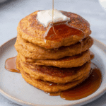 Stack of fluffy sweet potato pancakes topped with butter and drizzled with maple syrup on a white plate.