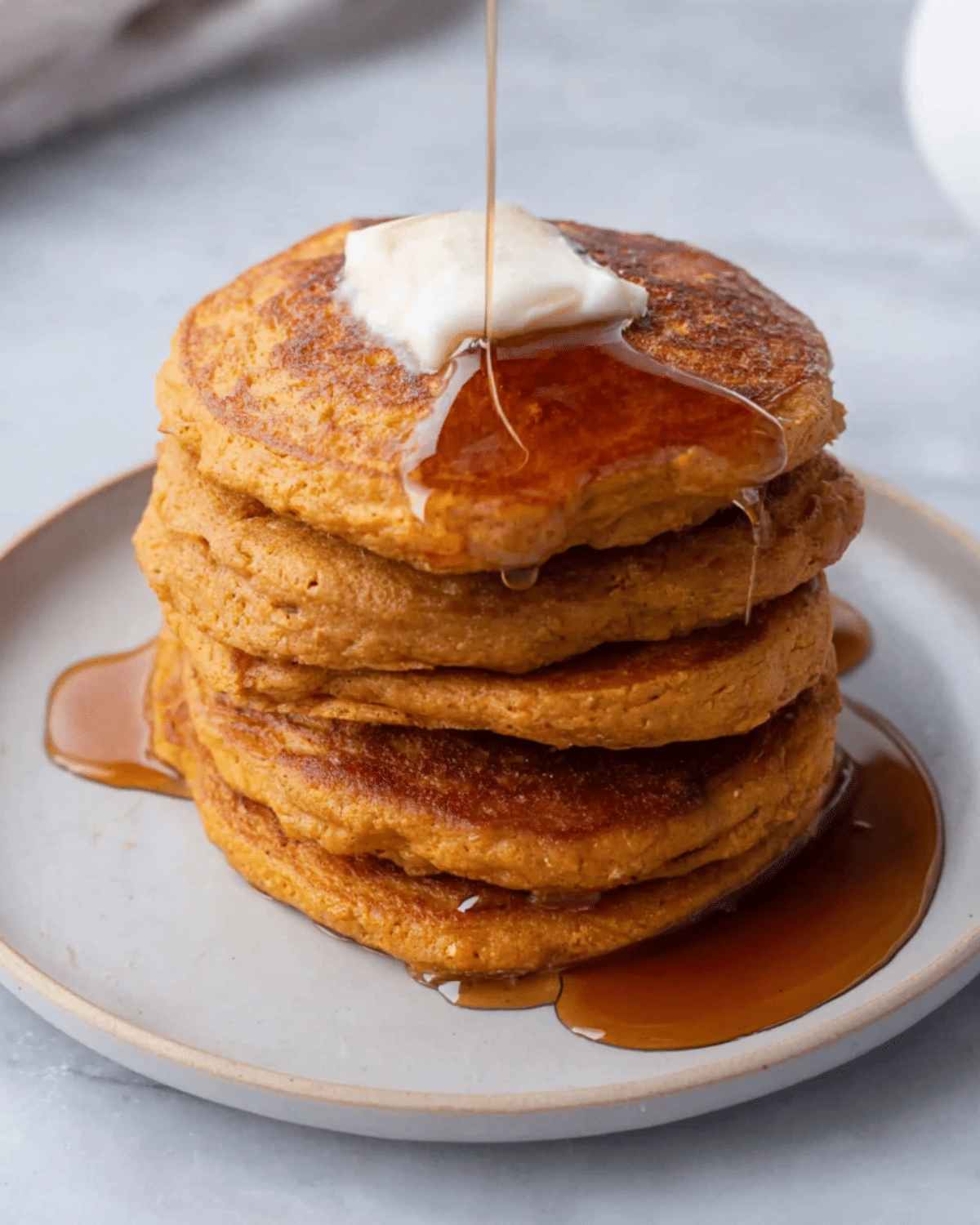 Stack of fluffy sweet potato pancakes topped with butter and drizzled with maple syrup on a white plate.