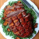 Fresh ham with cracklings, sliced and served on a white plate over a bed of arugula with a knife placed on the side.