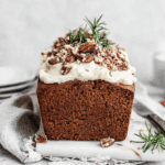Gingerbread loaf with frosting topped with chopped pecans and rosemary sprigs on a white surface.