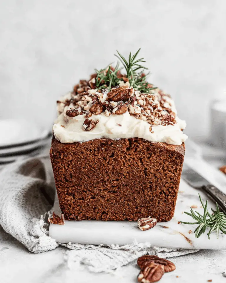 Gingerbread loaf with frosting topped with chopped pecans and rosemary sprigs on a white surface.