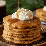 Stack of fluffy gingerbread pancakes topped with whipped cream, cinnamon, and maple syrup being poured over.