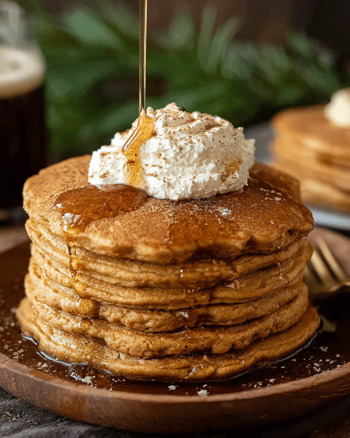 Stack of fluffy gingerbread pancakes topped with whipped cream, cinnamon, and maple syrup being poured over.