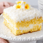 Slice of Hawaiian Wedding Cake topped with shredded coconut and yellow plumeria flowers.
