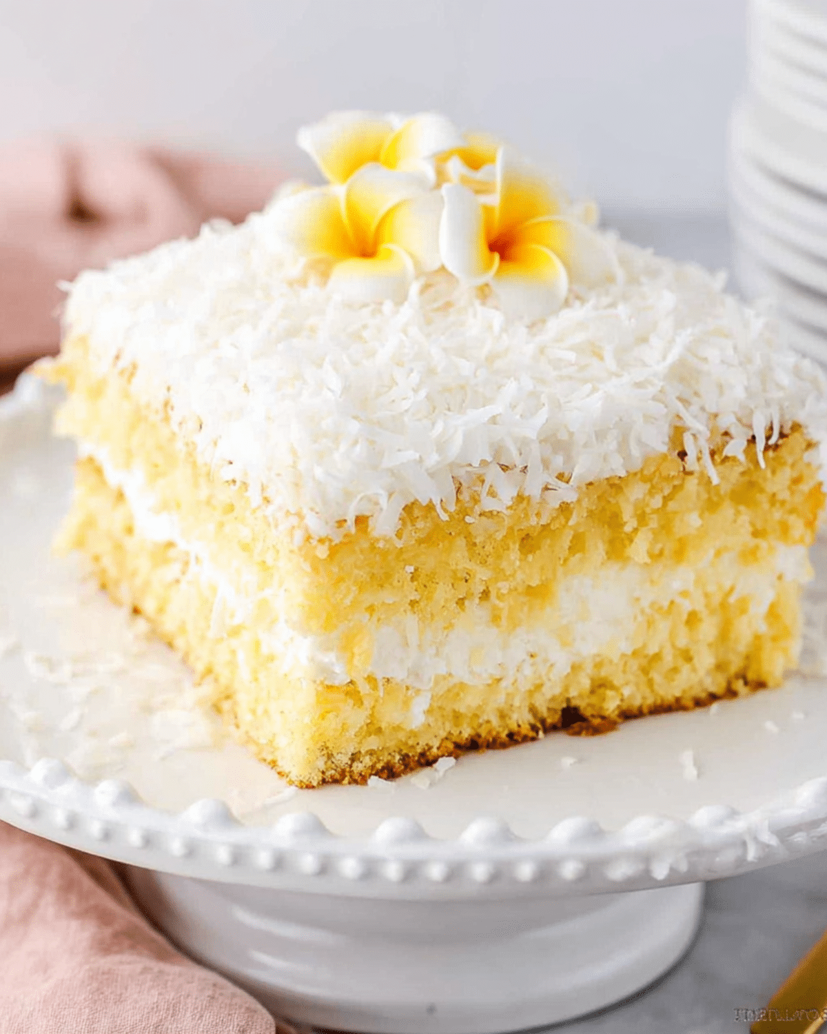 Slice of Hawaiian Wedding Cake topped with shredded coconut and yellow plumeria flowers.