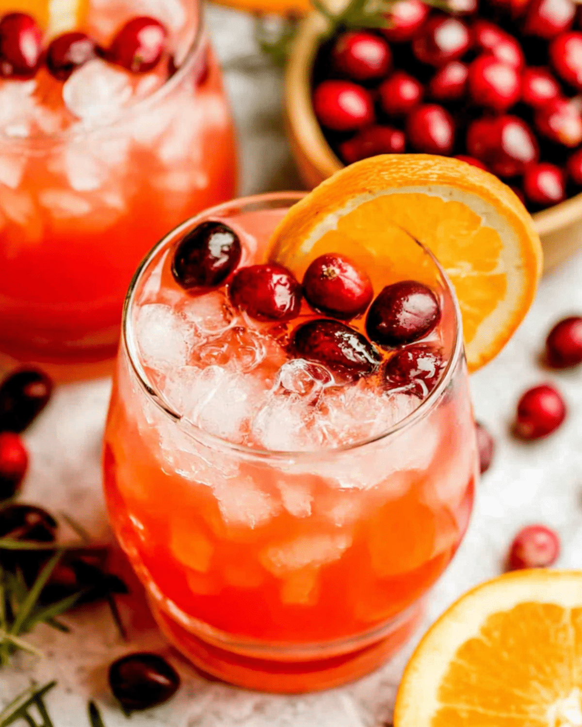A glass of festive holiday punch filled with ice, cranberries, and garnished with an orange slice, surrounded by fresh cranberries and citrus.
