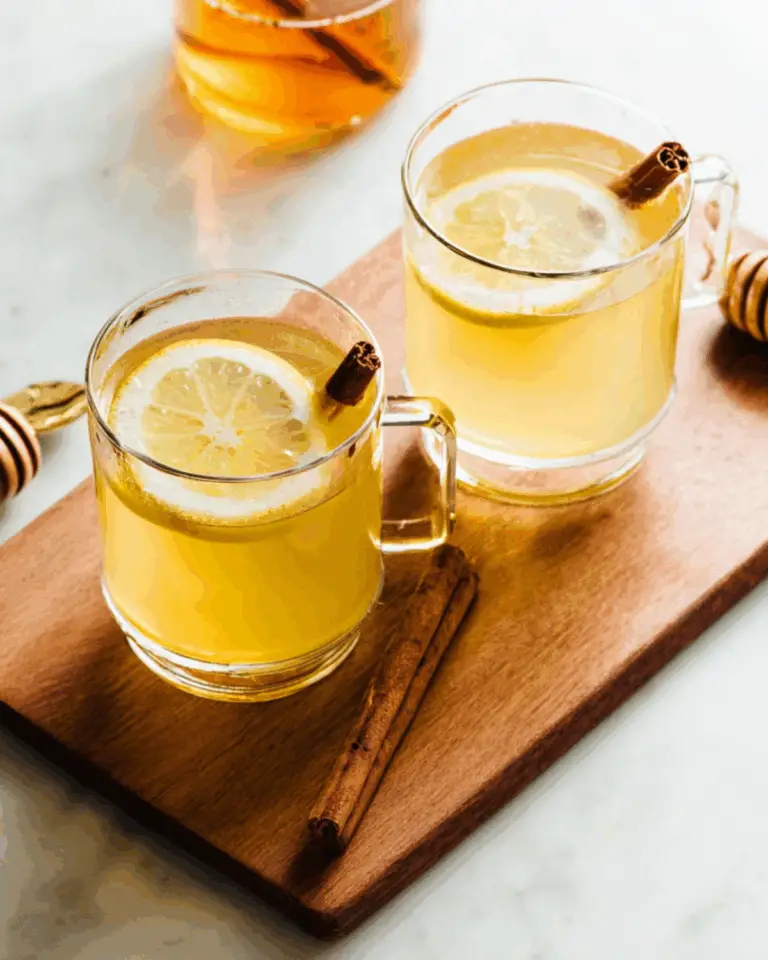 Two glass mugs of Hot Toddy (cocktail) garnished with lemon slices and cinnamon sticks, placed on a wooden board.