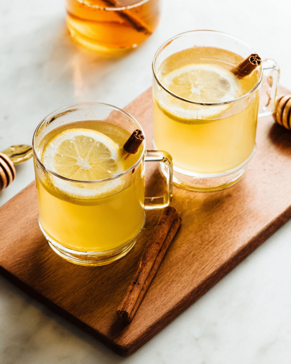 Two glass mugs of Hot Toddy (cocktail) garnished with lemon slices and cinnamon sticks, placed on a wooden board.