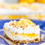 A slice of lemon lush dessert with layers of whipped cream, lemon pudding, and a nutty crust, garnished with chopped nuts and a lemon slice.