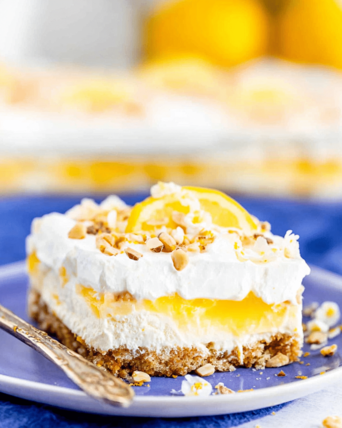 A slice of lemon lush dessert with layers of whipped cream, lemon pudding, and a nutty crust, garnished with chopped nuts and a lemon slice.