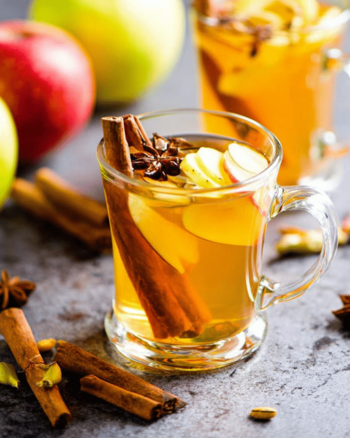 Glass mugs filled with mulled cider garnished with apple slices, cinnamon sticks, and star anise, surrounded by fresh apples and spices.