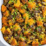 Close-up of a casserole dish filled with golden brown mushroom and herb stuffing, featuring toasted bread cubes, chopped mushrooms, celery, and fresh herbs.