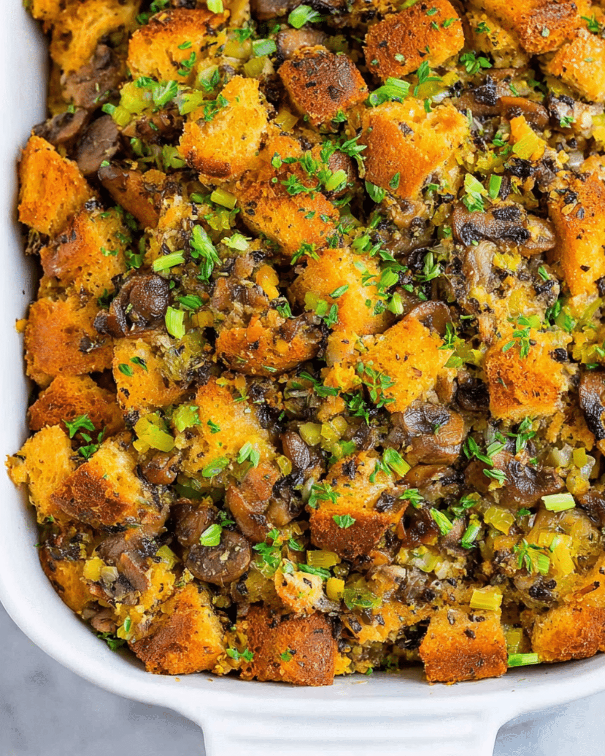 Close-up of a casserole dish filled with golden brown mushroom and herb stuffing, featuring toasted bread cubes, chopped mushrooms, celery, and fresh herbs.