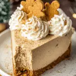A slice of no bake gingerbread cheesecake topped with whipped cream and mini gingerbread cookies on a white plate.