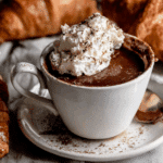 Cup of rich Paris Hot Chocolate topped with whipped cream and chocolate shavings, surrounded by croissants.