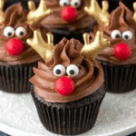 Chocolate reindeer cupcakes decorated with gold antlers, candy eyes, and red noses on a white plate.