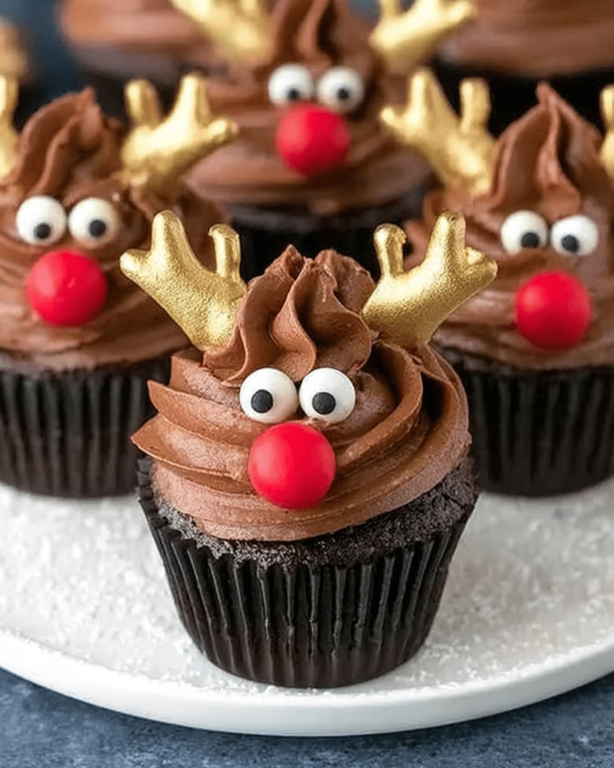Chocolate reindeer cupcakes decorated with gold antlers, candy eyes, and red noses on a white plate.