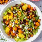 A vibrant roasted vegetable winter salad with arugula, chickpeas, Brussels sprouts, squash, red onions, and crumbled feta in a white bowl.