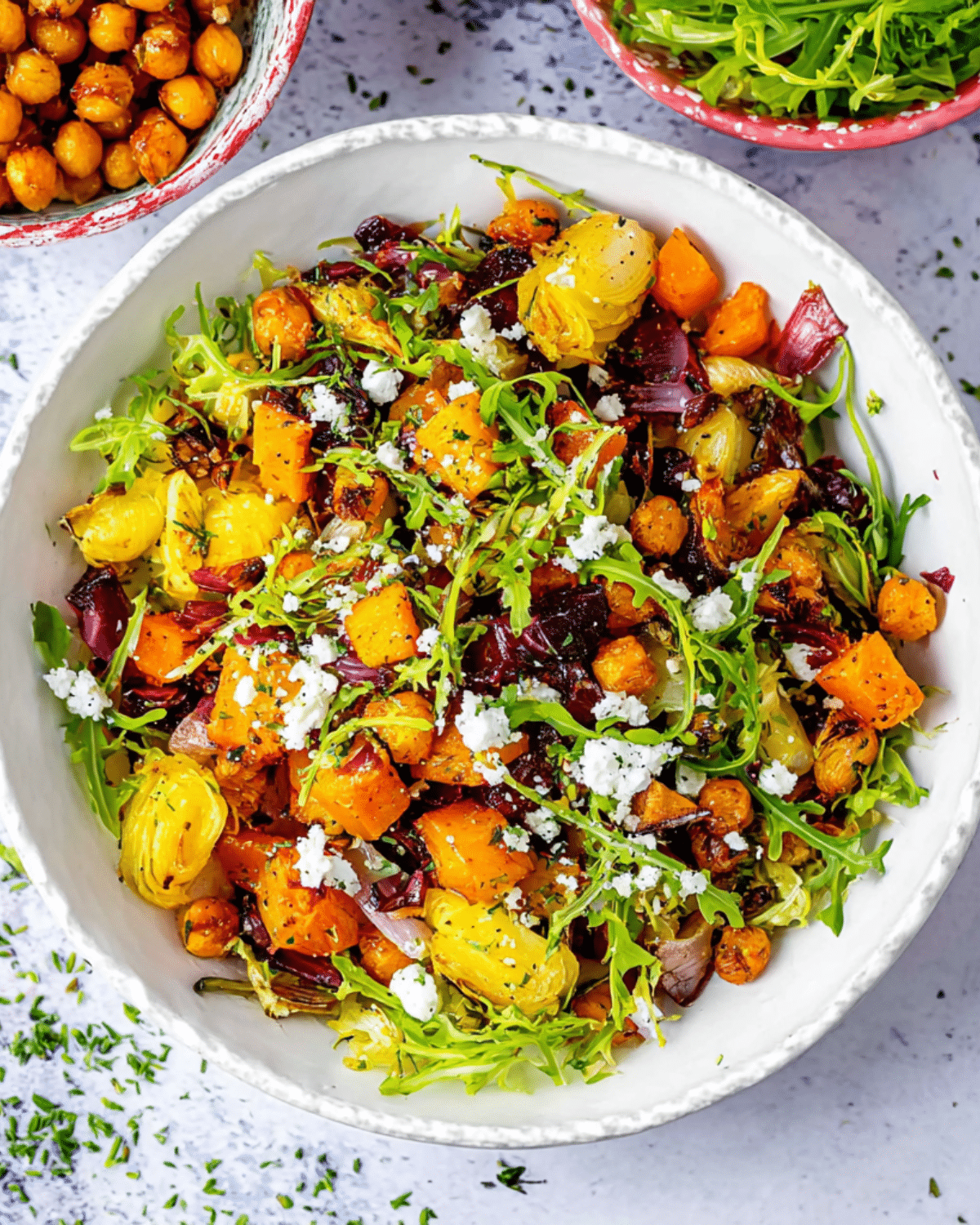 A vibrant roasted vegetable winter salad with arugula, chickpeas, Brussels sprouts, squash, red onions, and crumbled feta in a white bowl.