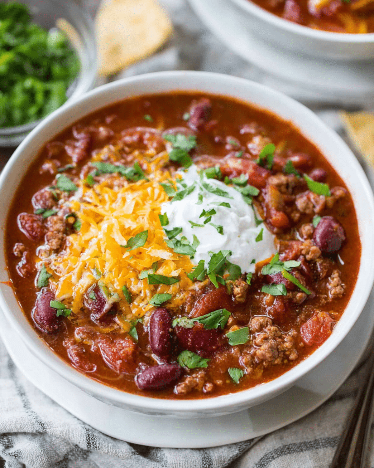 Bowl of slow cooker chili topped with shredded cheddar cheese, sour cream, and chopped parsley, served on a cloth napkin.