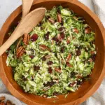 A wooden bowl filled with shredded Brussels sprouts salad, mixed with pecans, dried cranberries, and grated cheese, with wooden salad servers.