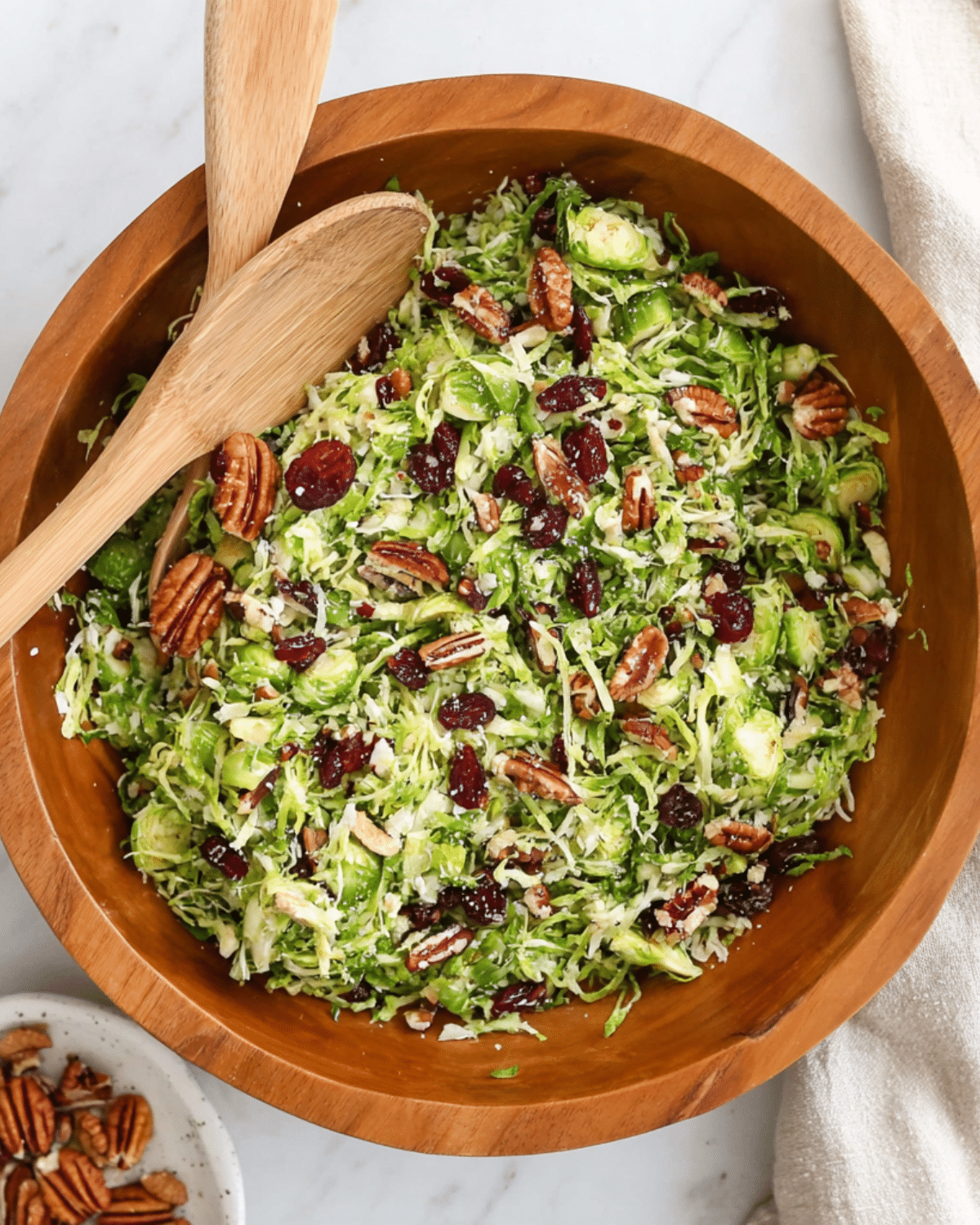 A wooden bowl filled with shredded Brussels sprouts salad, mixed with pecans, dried cranberries, and grated cheese, with wooden salad servers.