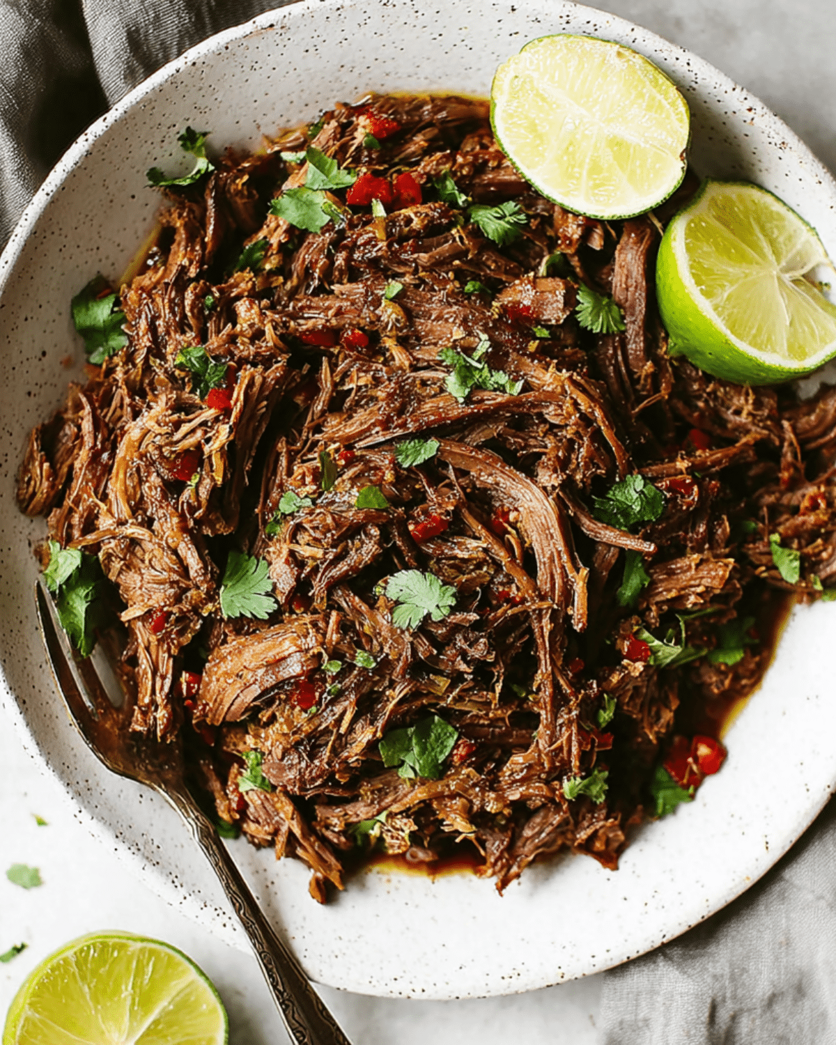 Slow cooker beef barbacoa served on a white plate, garnished with fresh cilantro and lime wedges.