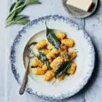 Squash gnocchi served on a plate with brown butter and crispy sage, topped with grated cheese.