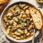 A hearty bowl of white bean mushroom stew garnished with fresh herbs and served with crusty bread.