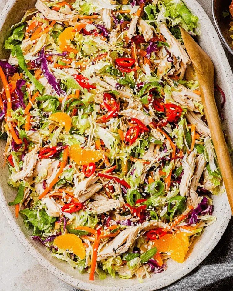 A vibrant bowl of Asian Chicken Salad with shredded chicken, lettuce, red cabbage, carrots, mandarin oranges, red chili slices, and sesame seeds.