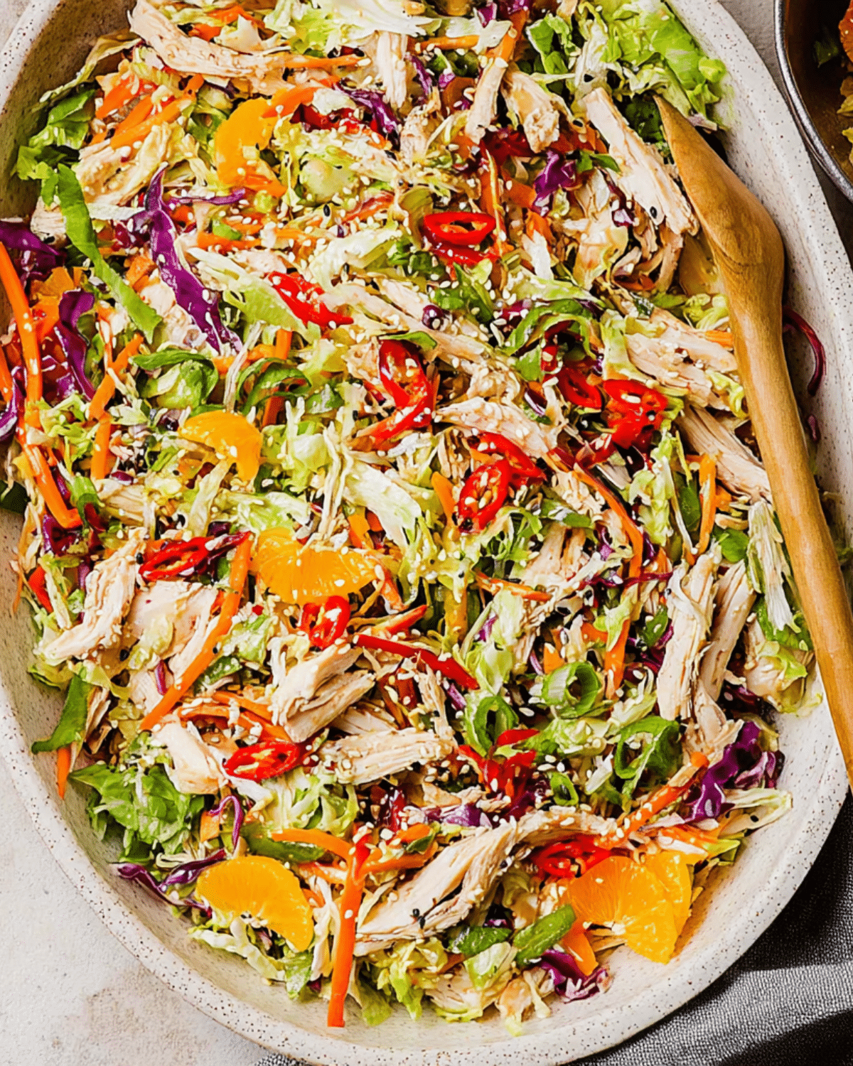 A vibrant bowl of Asian Chicken Salad with shredded chicken, lettuce, red cabbage, carrots, mandarin oranges, red chili slices, and sesame seeds.