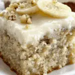 Moist slice of Banana Bread Cake with Cream Cheese Frosting, topped with banana slices and chopped nuts on a white plate.
